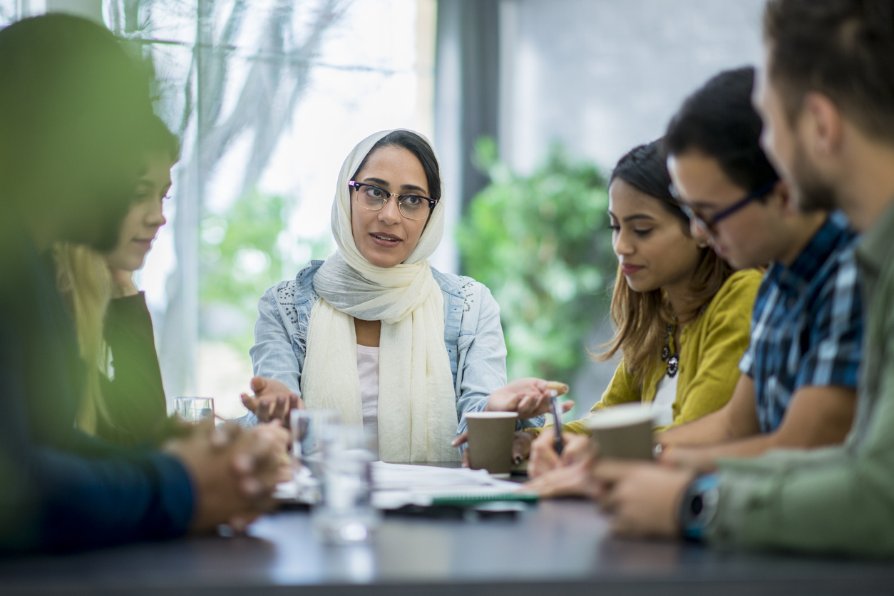 A multi-ethnic group of business people are indoors in an office boardroom. They are sitting around a table and making plans. A woman wearing a head scarf is explaining papers to her coworkers.