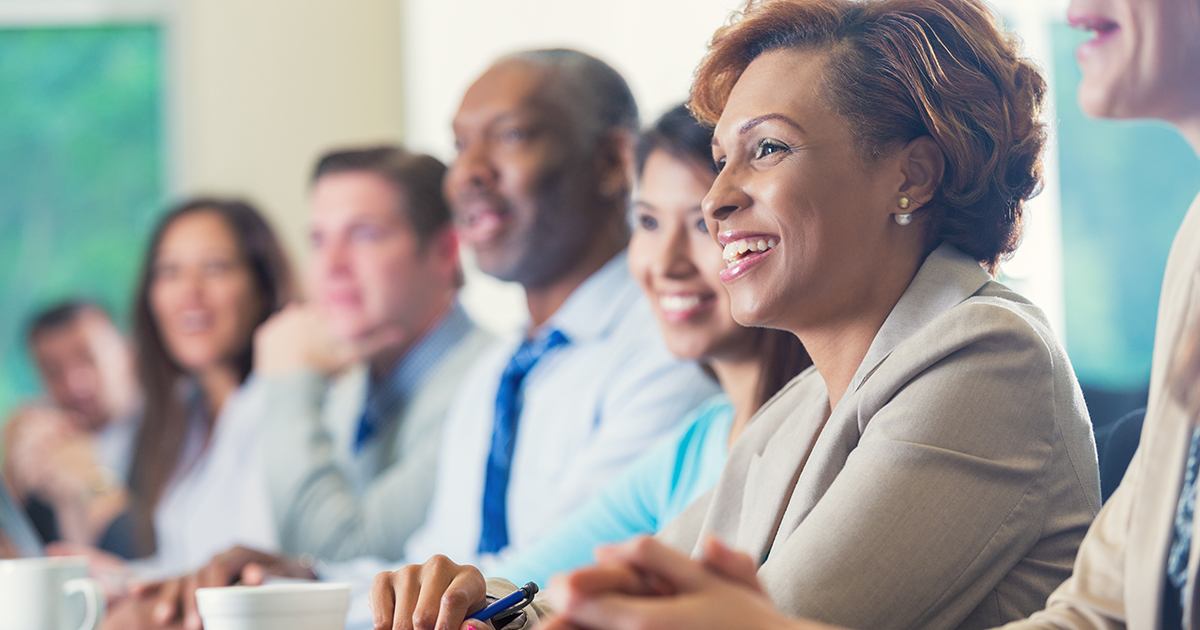 Mid adult African American businesswoman is smiling while attending business conference. She is listening to speaker in seminar while she sits in a row with other diverse professional businesspeople. Woman is wearing business casual clothing and is taking notes. She's drinking a cup of coffee.