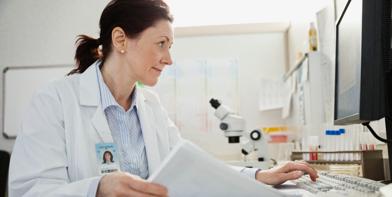 Doctor using computer while reviewing records at desk in clinic, individual, woman