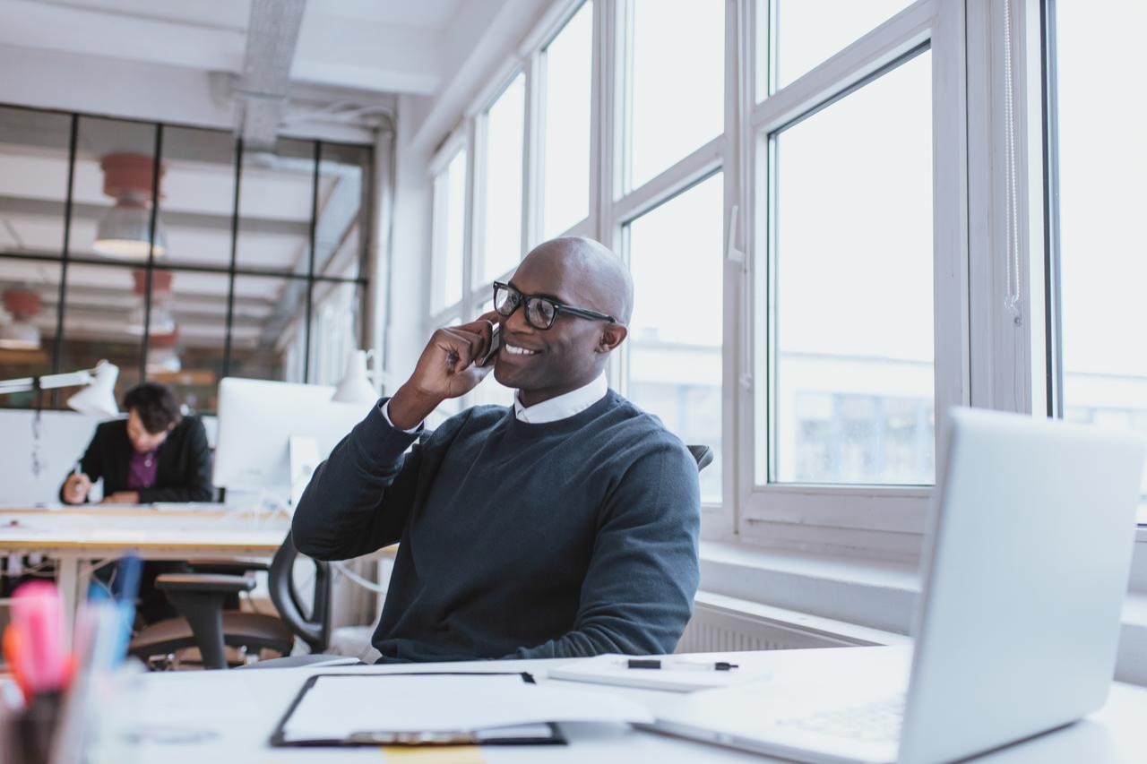 Young man talking on his mobile phone in office. African executive sitting at his desk with laptop